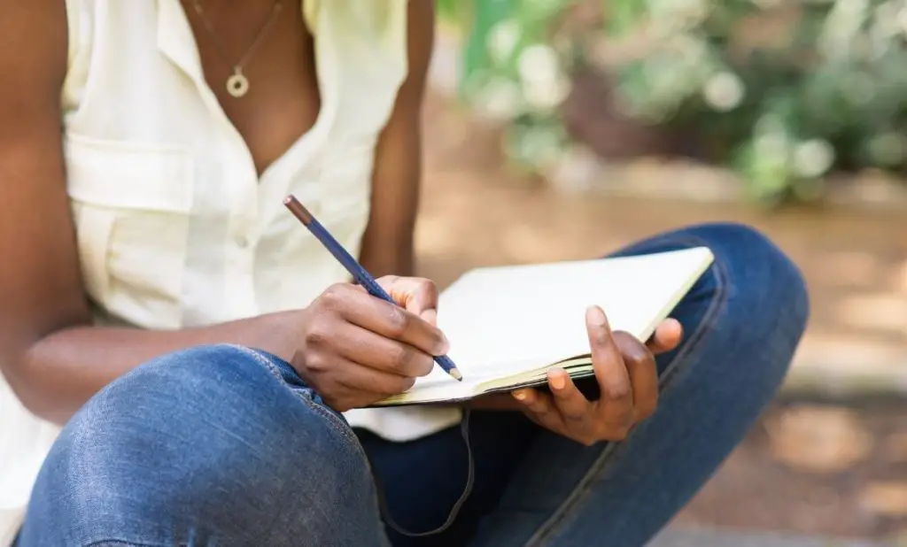 a woman sits cross-legged on the ground writing in a notebook about her finances. happytogetheranywhere.com