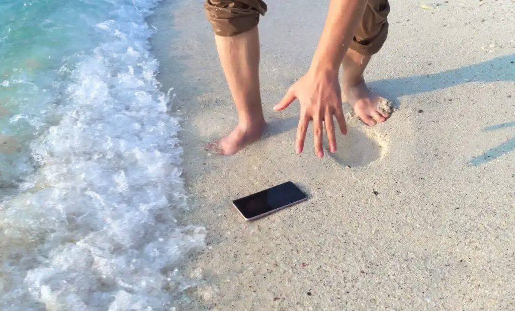 person wearing rolled up pants stands by the water on a sandy beach reaching for a dropped cell phone as a wave comes in. happytogetheranywhere.com