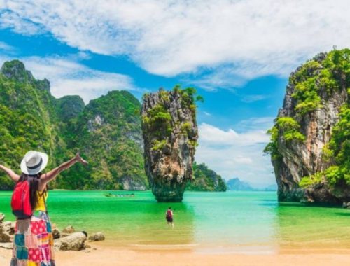 girl with backpack and bright skirt stands with arms spread on beach in thailand