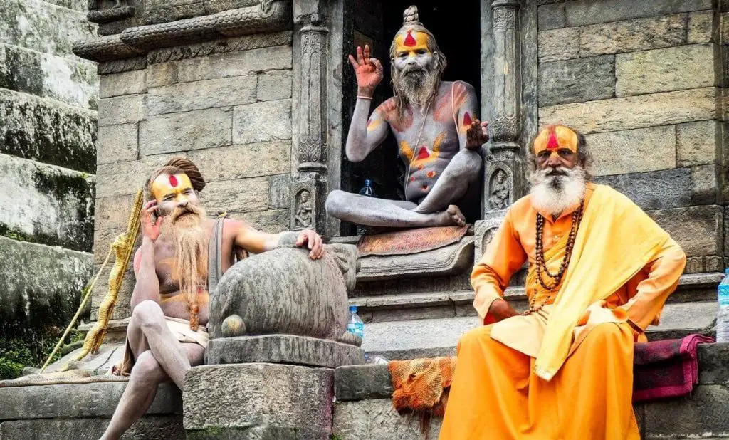 experience different cultures as a reason to travel; three holy men, one in a bright yellow robe, sit in front of a temple in kathmandu, india. happytogetheranywhere.com