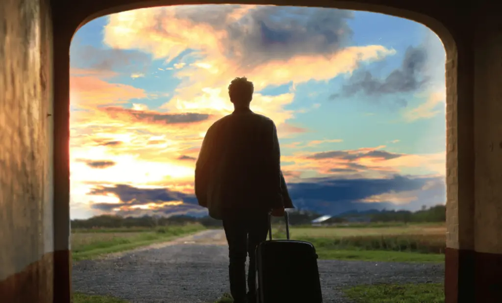 man walking out of archway towards a bright sunrise with a rolling suitcase as he embarks on a long trip. happytogetheranywhere.com