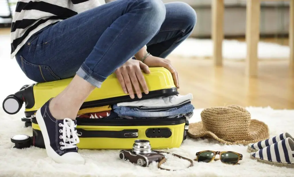 women in jeans and sneakers sitting on a bright yellow and black suitcase trying to close it, with sunglasses, a camera and a hat nearby. happytogetheranywhere.com
