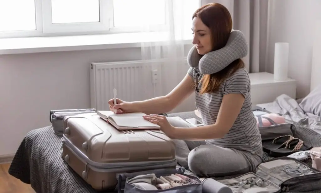 woman sitting on bed with a suitcase and piles of clothing wearing a neck pillow is writing in a notebook as she plans a packing list for southeast Asia. happytogetheranywhere.com