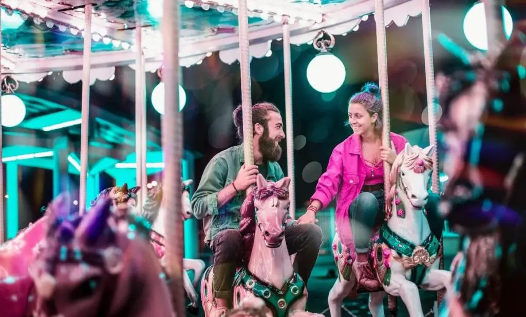 a man and woman holding hands and smiling at each other as they ride on horses on a bright carousel at night. happytogetheranywhere.com