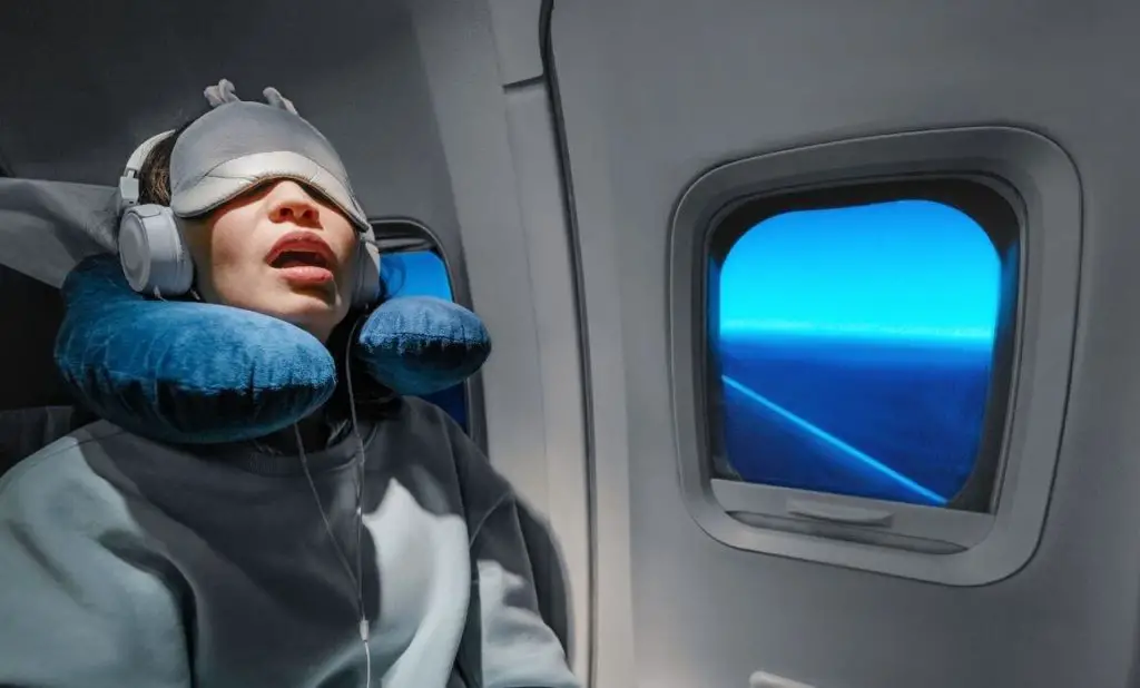 person sleeping on plane with open mouth, wearing a neck pillow, eye mask, and noise cancelling earphones. happytogetheranywhere.com
