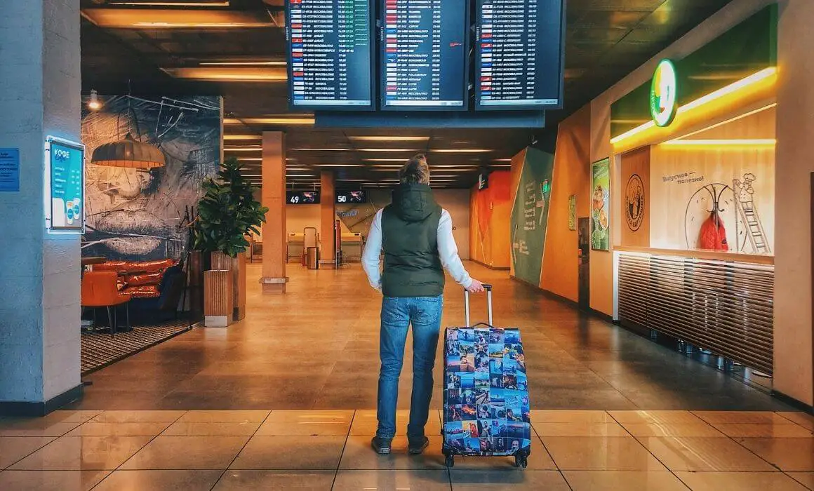 top reasons to travel man looking at airport departure screen with blue suitcase